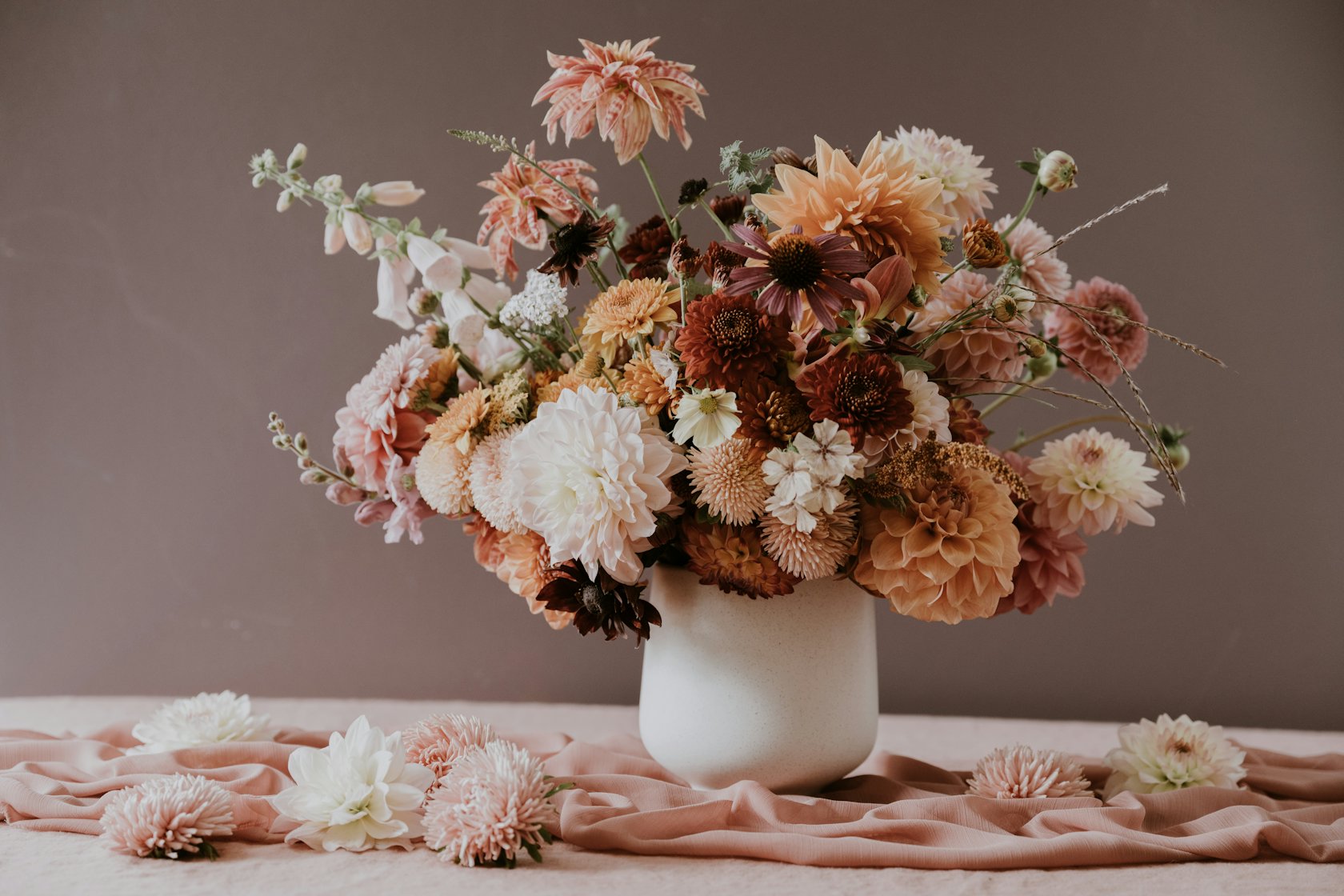A loose arrangement of peach and rust dahlias, foxglove and scabiosa in a cream stoneware vase on blush linen, against a mauve wall
