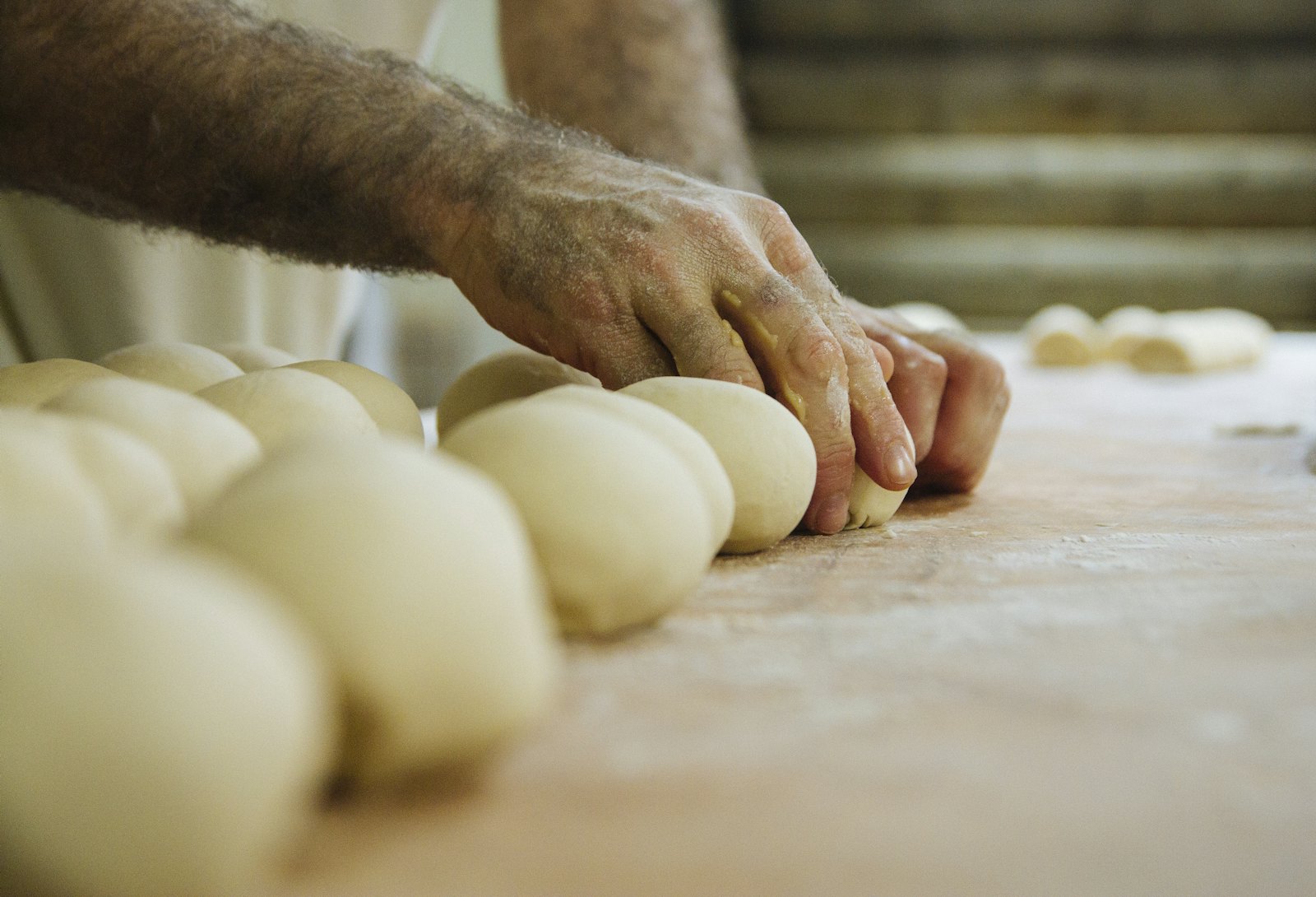 A baker's floured hands shaping rows of soft dough balls on a wooden bench, ovens blurred behind.