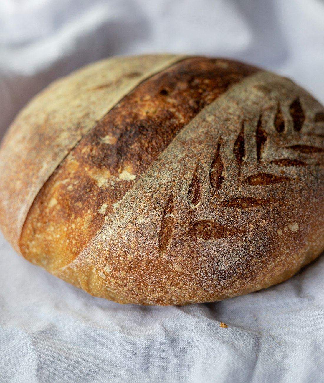 Close-up of a country sourdough loaf, scored with a wheat-ear pattern and dusted with flour, resting on white linen.