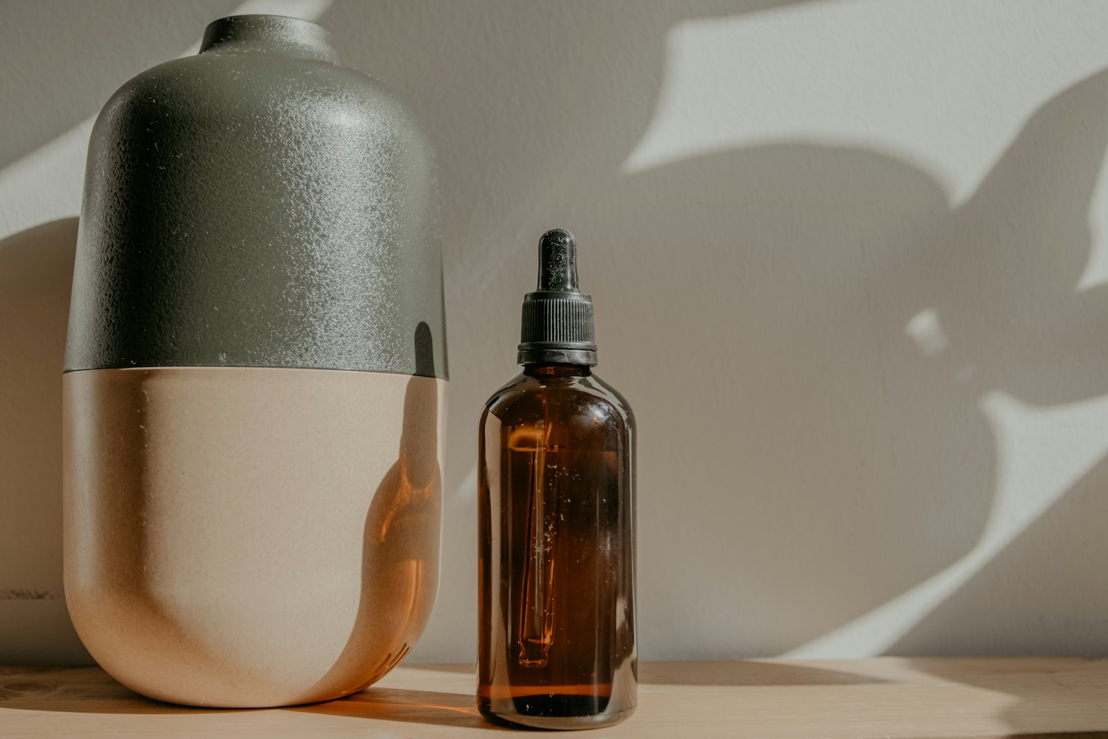 Amber glass dropper bottle beside a stoneware vase on a wooden shelf, palm leaf shadows falling across a warm plaster wall